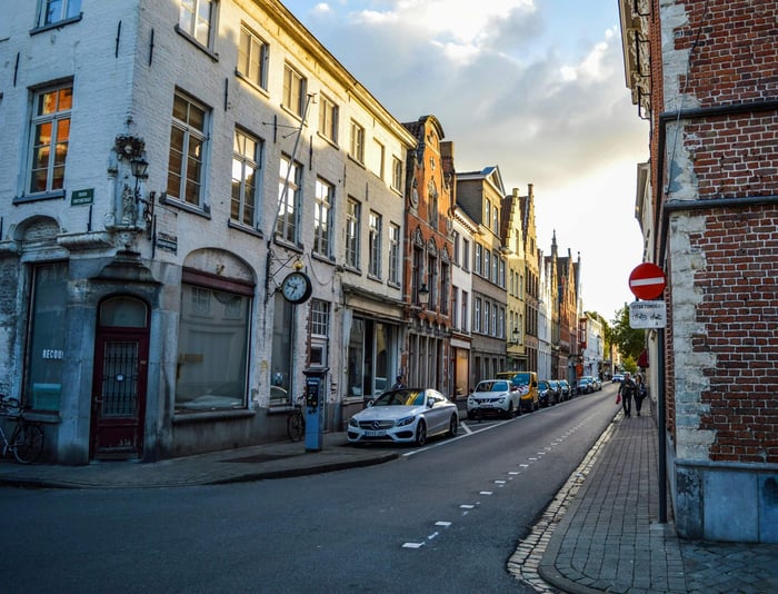 Low angle of asphalt road with parked vehicles near aged residential buildings in historical district of Bruges in sunny morning