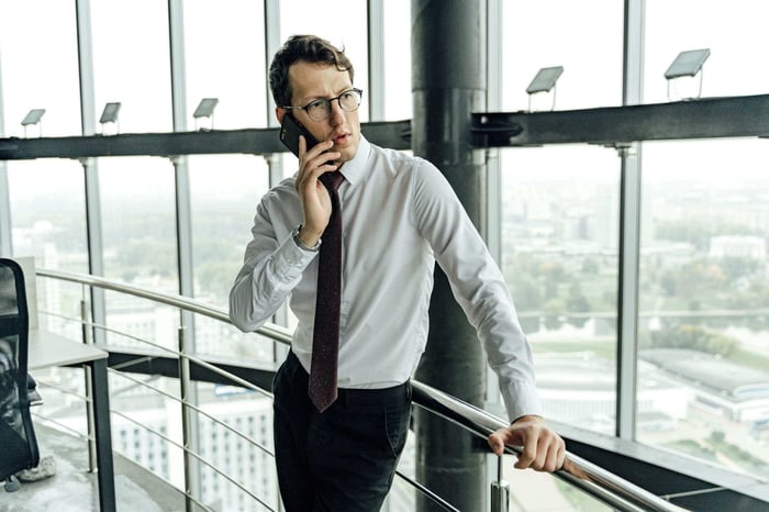 Young professional in a white shirt and tie making a phone call in a high-rise office with city view.