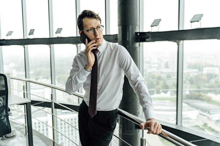 Young professional in a white shirt and tie making a phone call in a high-rise office with city view.