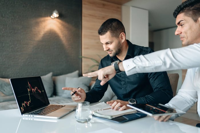 Two businessmen reviewing financial data on a laptop indoors, analyzing market trends.