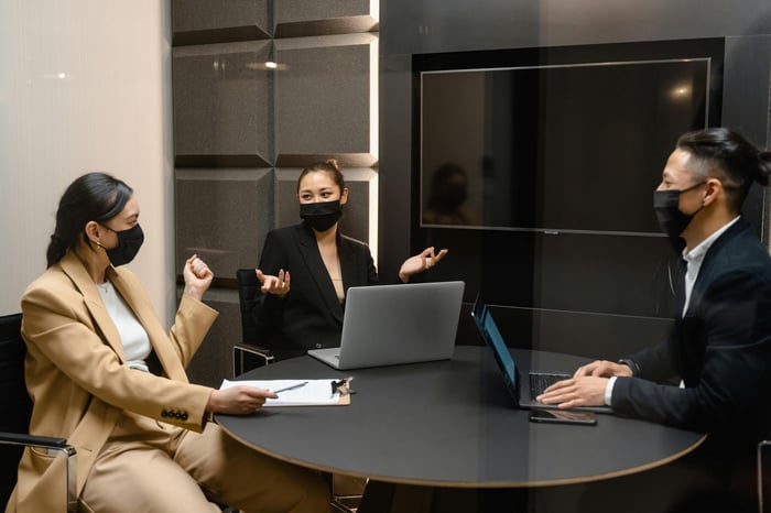 Three business professionals wear face masks during a meeting, collaborating around a round table indoors.