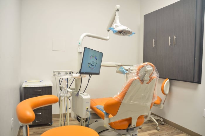 Modern dental exam room with orange patient chair, overhead exam light, monitor, and clean cabinetry.