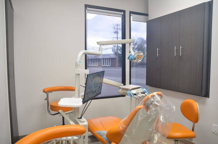Modern dental exam room with orange chair, overhead light, monitor, and clean cabinetry beside large windows.