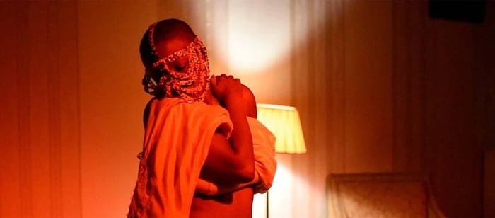 An image of Jennifer Hagre standing in a darkened room in front of a lamp. Jennifer holds a sheet on their arms and is looking down and to the right.