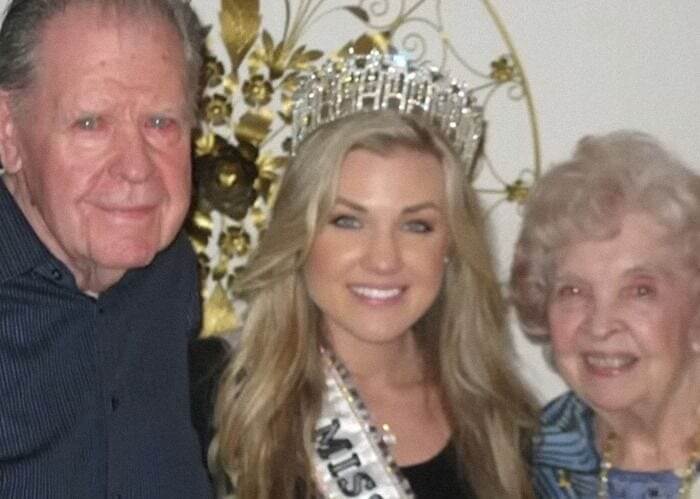 Erika Kirk wearing a crown and sash, posing with two elderly individuals in an indoor setting.