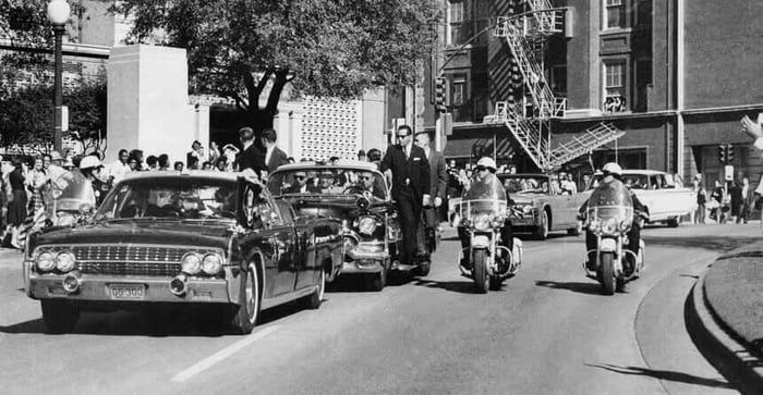 The motorcade proceeds along Elm Street past the Texas School Book Depository, shortly after Kennedy was shot. Looking through the limo's windshield in the foreground, Kennedy appears to raise his hand toward his head after being shot. The first lady holds his forearm in an effort to aid him. The motorcade proceeds along Elm Street past the Texas School Book Depository, shortly after Kennedy was shot. Looking through the limo's windshield in the foreground, Kennedy appears to raise his hand toward his head after being shot. The first lady holds his forearm in an effort to aid him.