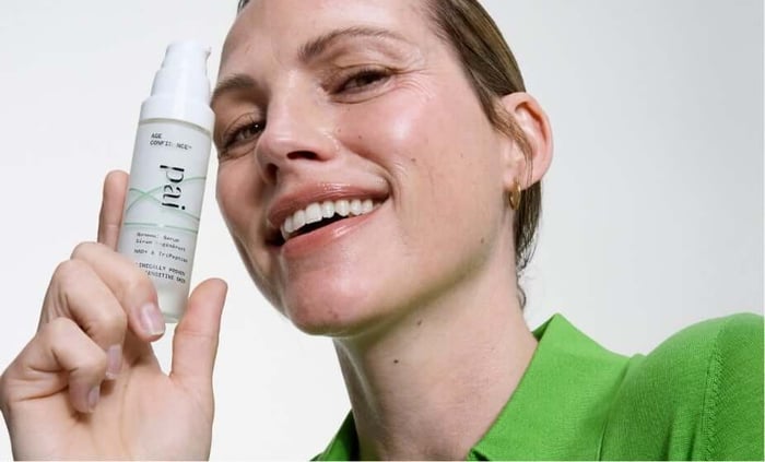 Smiling woman in green top holding Pai Age Confidence NAD+ Serum, showing the white pump bottle with minimalist packaging