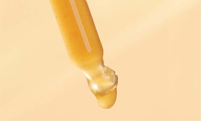 Golden honey-textured self tan drops being dispensed from dropper against warm cream background