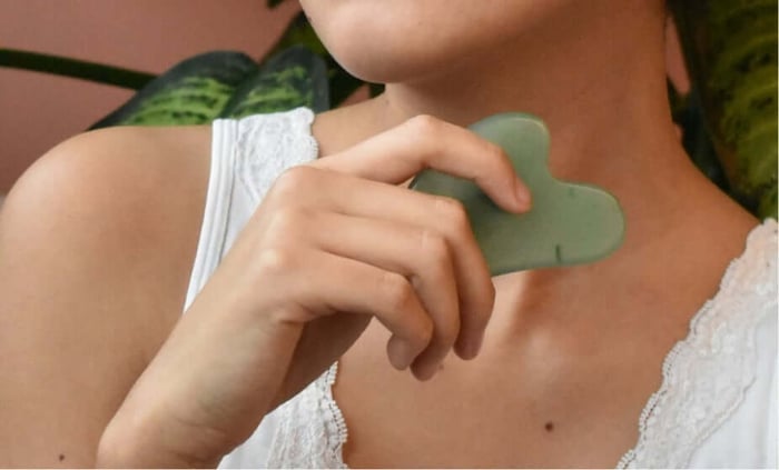Woman using green gua sha stone for facial massage on neck with plants in background
