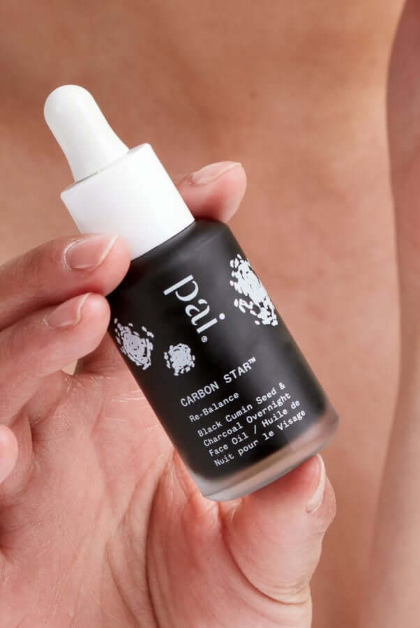 Hand holding Pai Carbon Star black cumin seed face oil with white dropper against warm background