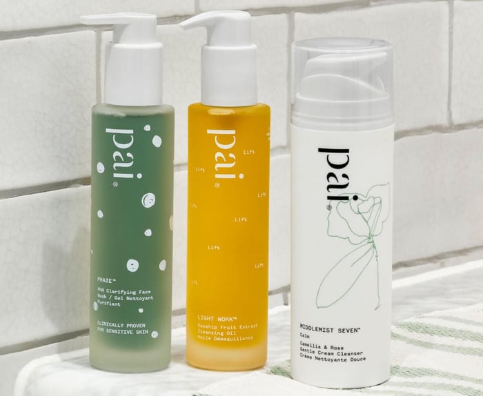 Three Pai Skincare cleansers on white bathroom tiles - green Phaze gel cleanser for sensitive skin, golden Light Work rosehip cleansing oil, and white Middlemist Seven cream cleanser