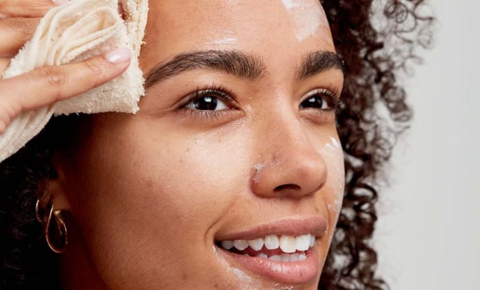 Woman with curly hair gently patting face dry with cream muslin cloth, showcasing healthy glowing skin after cleansing routine