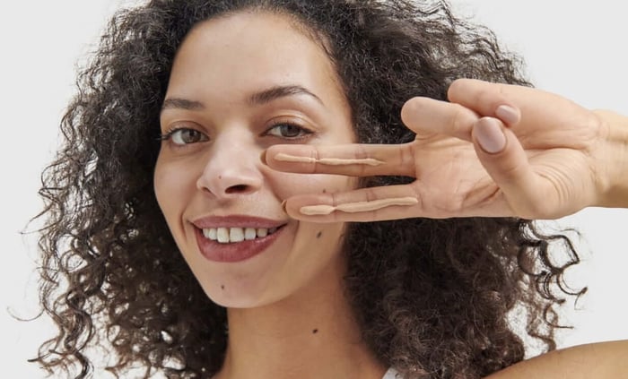 Woman with curly hair making a peace sign while smiling, showing healthy glowing skin wearing sunscreen