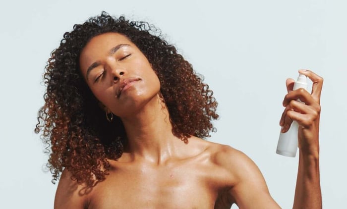 Woman with curly hair spritzing Century Flower face mist with eyes closed, demonstrating hydrating skincare for skin barrier protection
