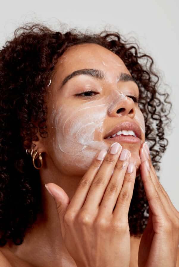 Woman with curly hair gently massaging white cream cleanser into her face with both hands