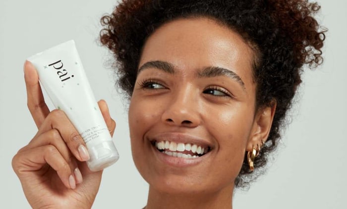 Happy woman with curly hair holding Pai Curtain Call brightening face mask, showcasing radiant glowing skin