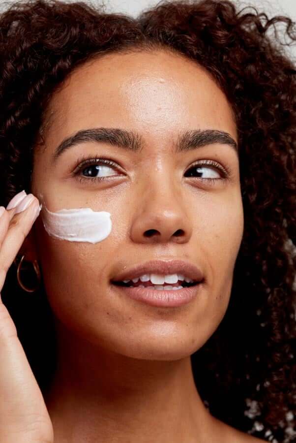 Woman with curly brown hair applying white face moisturiser to her cheek with fingertips