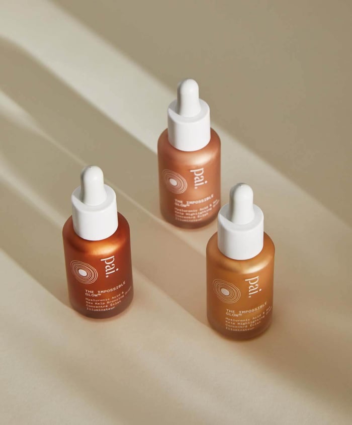 Three Pai Impossible Glow drops in warm amber-toned bottles with white droppers arranged on beige surface with dramatic lighting