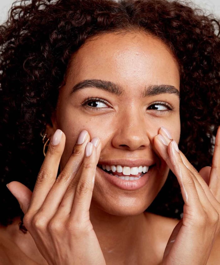 Woman with curly hair smiling while gently applying skincare to face, demonstrating gentle care for sensitive skin