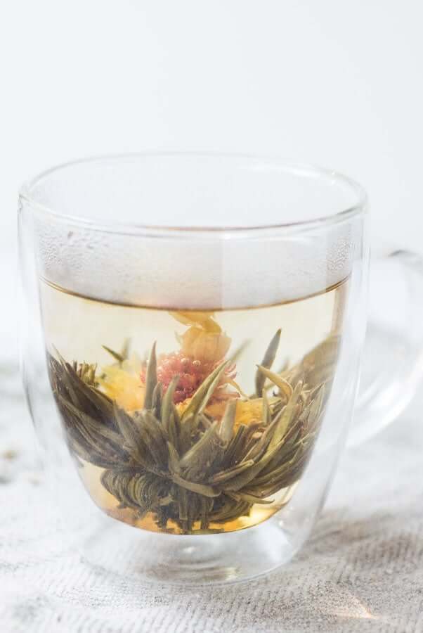 Blooming herbal tea with green leaves and colorful flower petals steeping in clear glass cup
