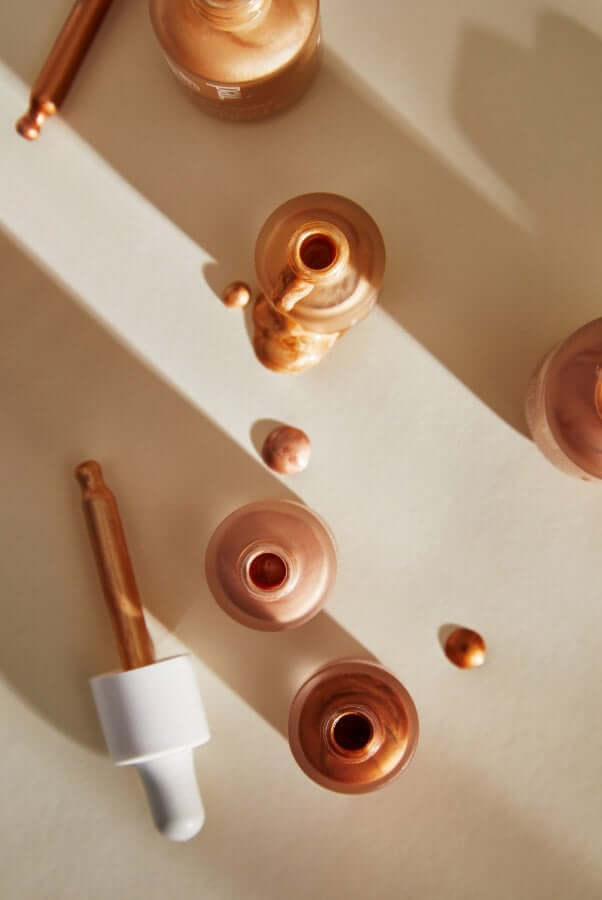 Copper-toned glow drops with rose gold packaging arranged in warm sunlight, showing reflective mica particles