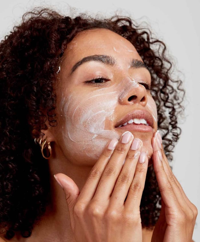Woman with curly hair applying gentle cleanser to her face with both hands, eyes closed in relaxation