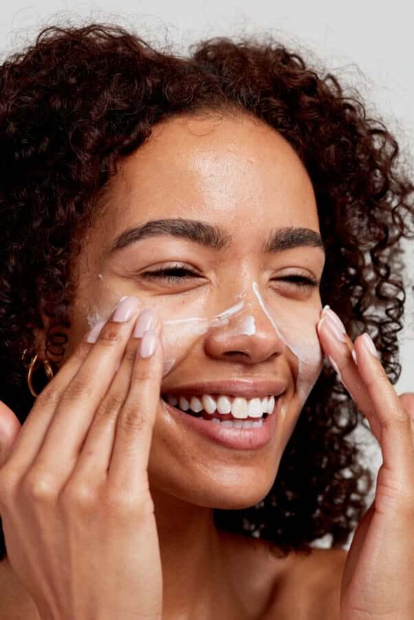 Smiling woman with curly hair applying white cream cleanser to her face with both hands