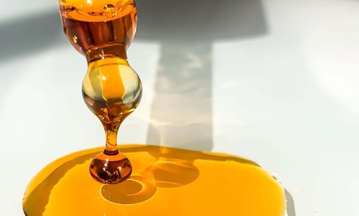 Golden honey-colored oil dripping from wooden spoon creating ripples in amber pool below