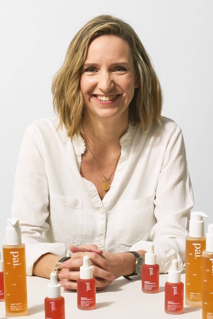 Sarah Brown, Pai Skincare founder and CEO, smiling at camera surrounded by orange and red Pai skincare products
