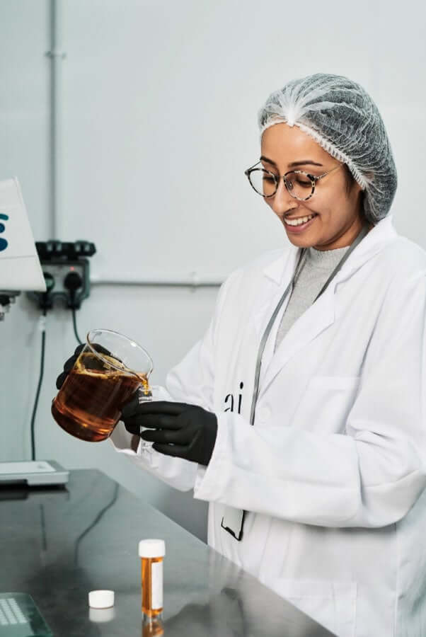 Female scientist in lab coat examining golden-colored skincare formula in glass beaker