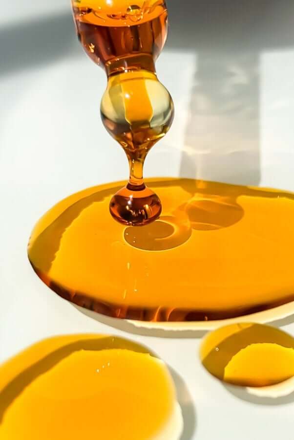 Golden amber rosehip oil dropping from glass dropper into white ceramic dish, showing pure oil texture