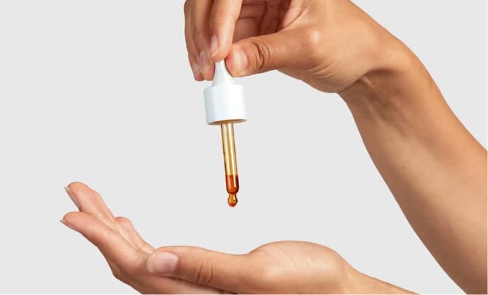Hand applying golden facial oil drops from white dropper bottle onto palm for dry skin care