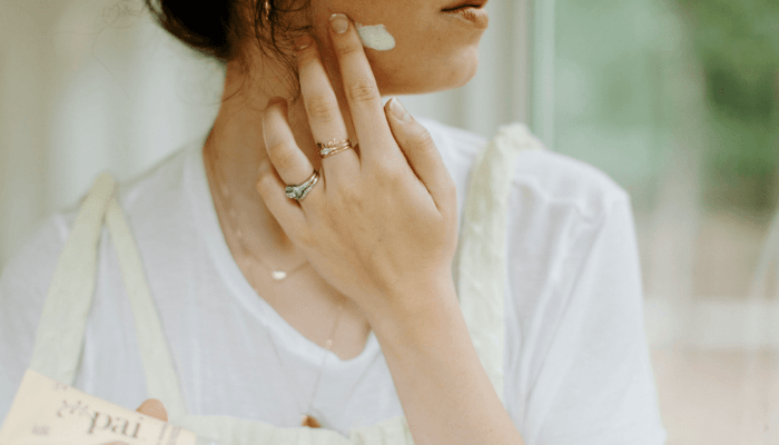 Woman applying white sunscreen cream to her face with fingertips, wearing delicate rings and white clothing