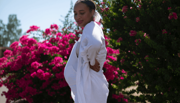 Pregnant woman in white robe outdoors surrounded by vibrant pink bougainvillea flowers, showcasing natural pregnancy glow