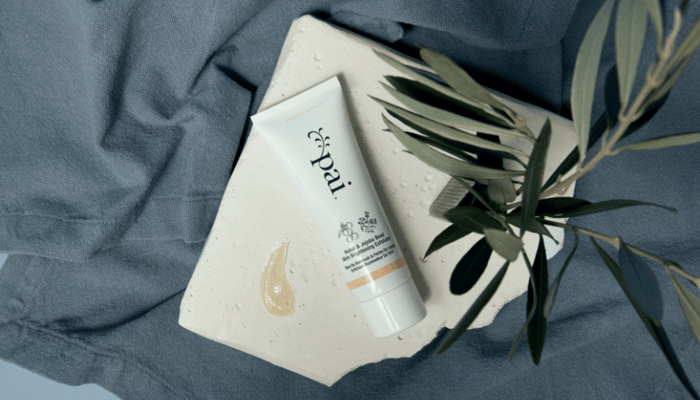 Pai Kukui & Jojoba Bead Skin Brightening Exfoliator tube with golden oil texture sample on white surface, styled with eucalyptus leaves