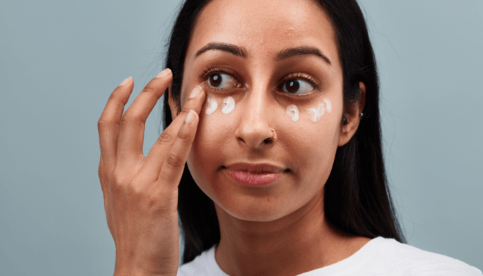 Woman applying white eye cream under both eyes with fingertip on clean grey background