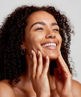 Woman with curly hair smiling while gently touching her glowing face, demonstrating healthy skin from rosehip oil benefits
