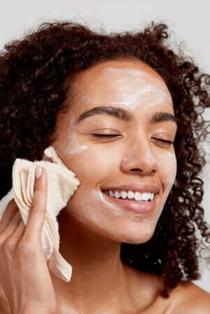 Woman with curly hair gently removing gentle exfoliator with muslin cloth, eyes closed and smiling