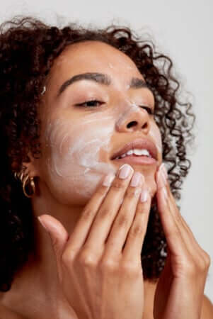 Woman with curly hair applying white facial cleanser to her face with both hands during morning skincare routine