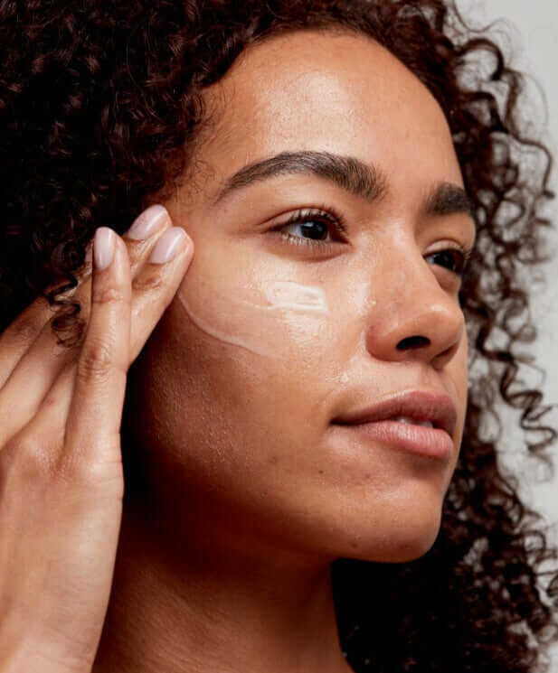 Woman with curly hair gently applying face moisturizer to her cheek with fingertips, demonstrating proper morning skincare routine technique