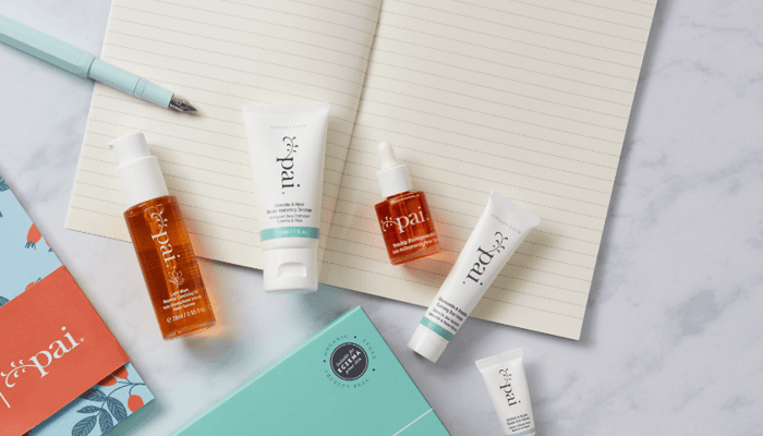 Pai Skincare rosacea products flatlay including orange Rosehip BioRegenerate Oil, white Instant Calm Redness Serum, and gentle cleanser tubes on marble surface with mint green notebook