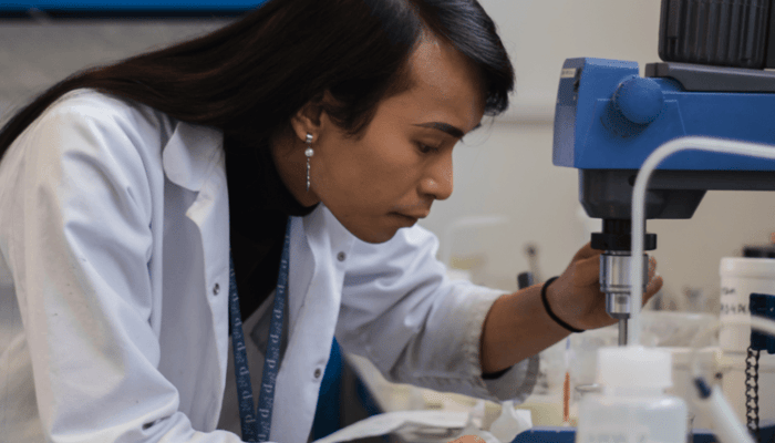 Female chemist in white lab coat using microscope while developing hand sanitiser formula in sterile laboratory