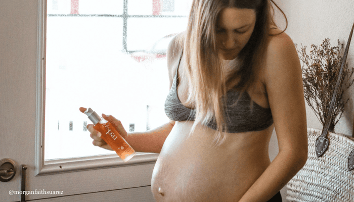 Pregnant woman in grey bra holding orange Pomegranate & Pumpkin Seed Stretch Mark System bottle by bright window