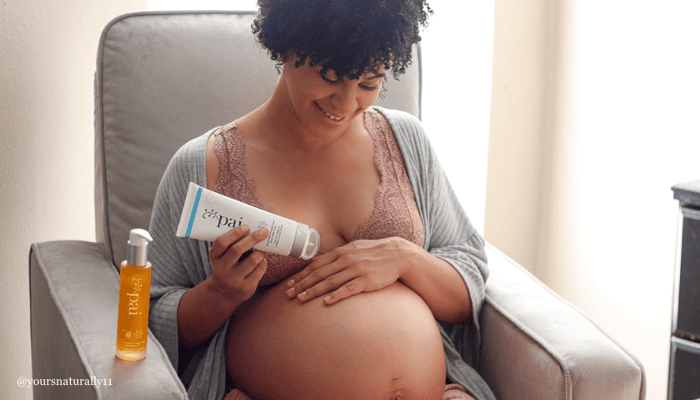 Pregnant woman with curly hair applying Pai Pomegranate & Pumpkin Seed Stretch Mark System to her belly while sitting in cozy grey chair