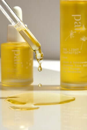 Golden rosehip oil dropping from pipette into small puddle, with Pai Rosehip BioRegenerate Oil bottle and packaging in background