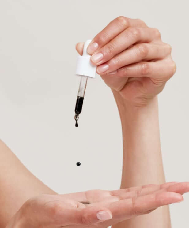 Hand dispensing dark oil serum from glass dropper bottle into palm for face application
