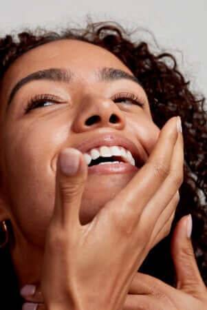 Woman with glowing combination skin touching her face and smiling, showcasing healthy complexion with natural curls