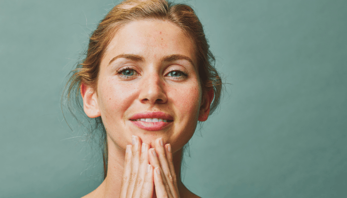 Woman with glowing skin gently applying facial oil with fingertips against sage green background