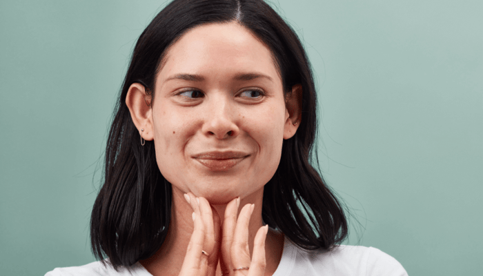 Woman with dark hair touching her neck and face, demonstrating skincare routine on sage green background