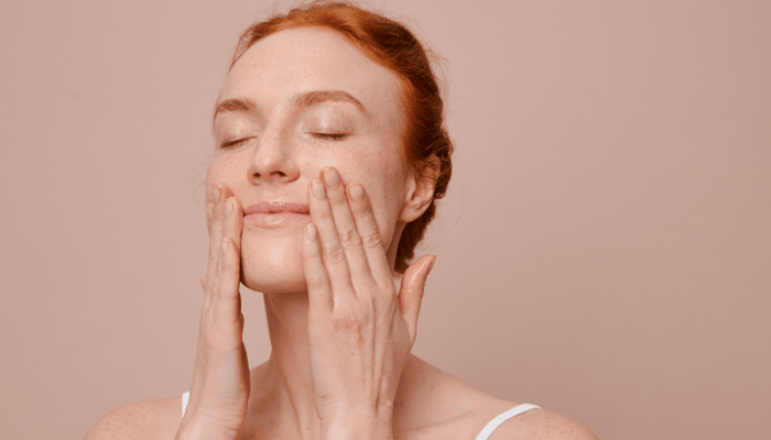 Woman with red hair gently cupping face with both hands, eyes closed in peaceful skincare routine moment